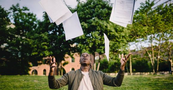 Job Searching, Graduate - Overjoyed African American graduate tossing copies of resumes in air after learning news about successfully getting job while sitting in green park with laptop